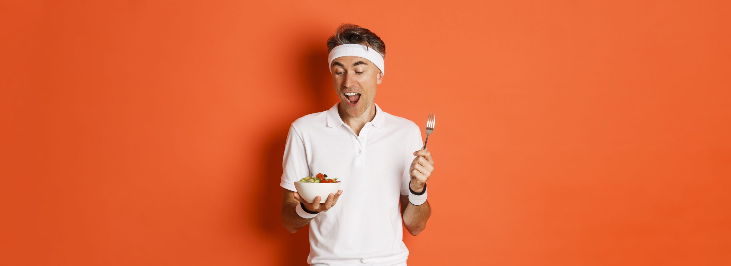 Concept of sport, fitness and lifestyle. Portrait of joyful middle-aged guy in workout uniform, holding fork and salad, eating healthy food, standing over orange background